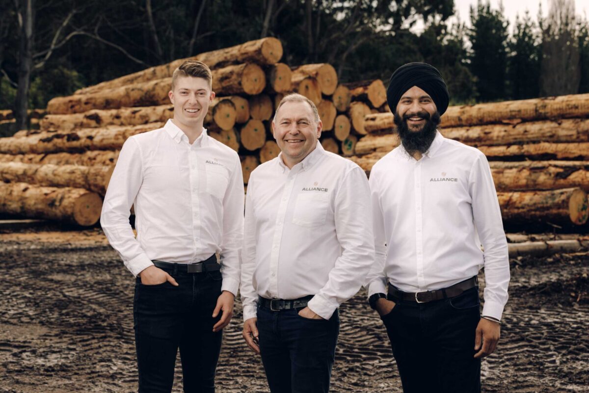 Three men in alliance woodware uniforms stand in front of piles of cut and trimmed pine logs