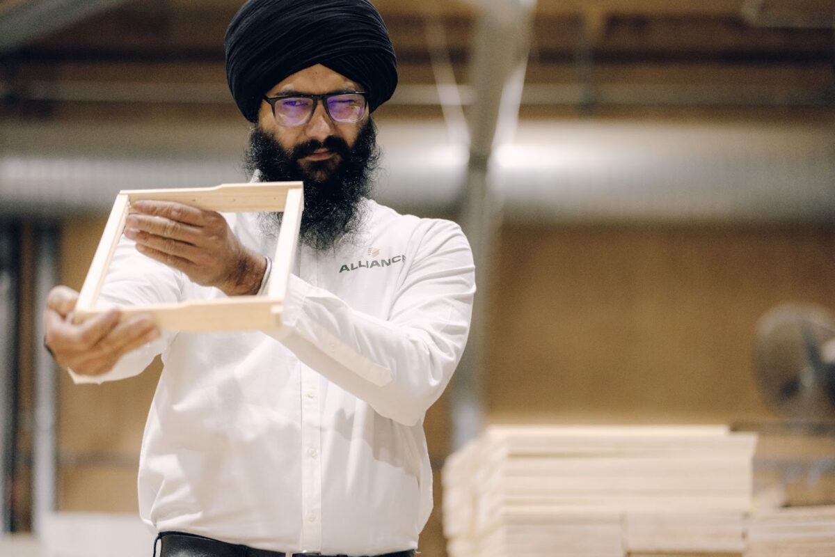 A team member in an Alliance Woodware shirt examines a handcrafted wooden frame in a workshop, holding it up to check its quality and alignment.