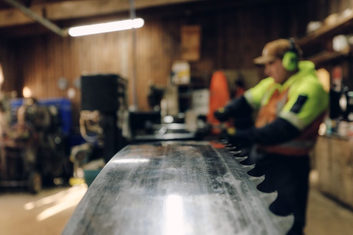 A forestry worker in high-visibility gear examines a large circular saw blade in a workshop, with the blade in focus and the workshop environment softly blurred behind.